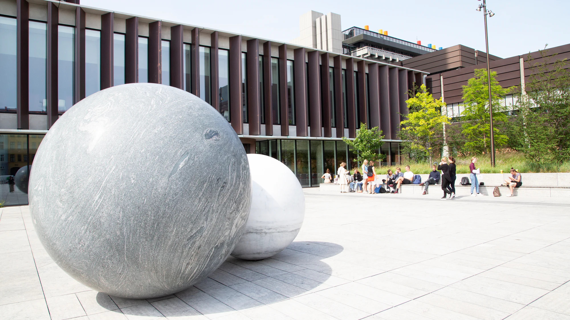 University of Copenhagen students gather in the sunlit courtyard at the Mærsk Tower on North Campus, with the building's deep vertical facade and two stone spheres in view. Photograph by Anna Palmehag for News Øresund, cropped from the original, CC BY 2.0.