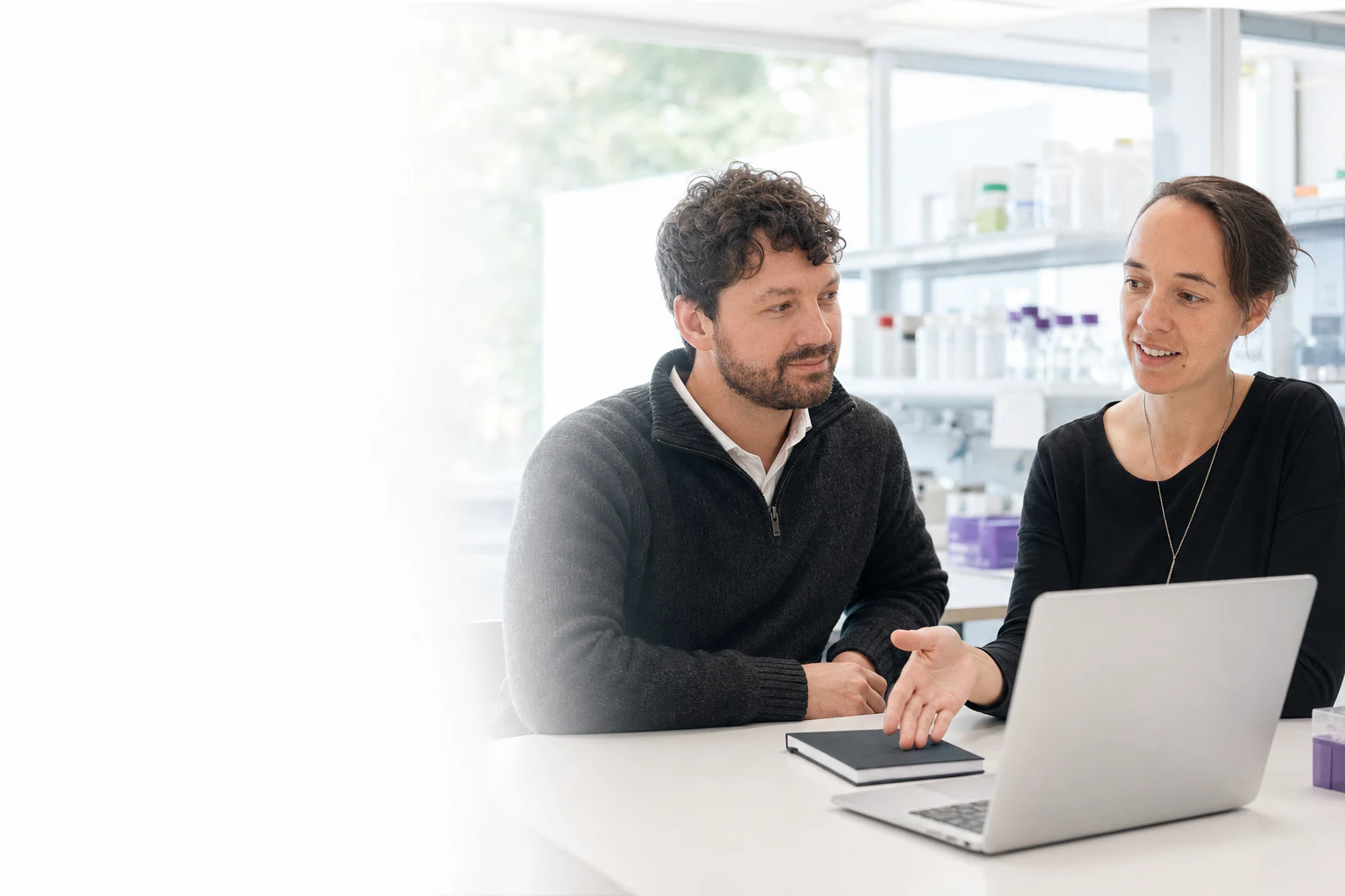 Two Peptone team members discussing scientific work beside a laptop in a bright laboratory. Illustrative generated image based on Peptone team portraits.